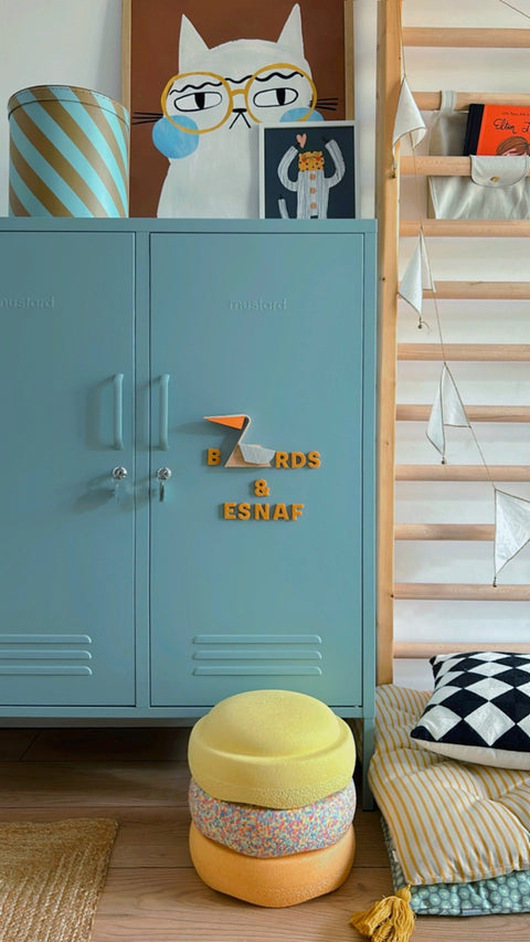 A styled interior scene showing a light blue metal locker with decorative magnets spelling 'BOARDS & ESNAF', a striped lamp, framed art, a wooden ladder shelf, and a round yellow-and-orange pouf on a wood floor.