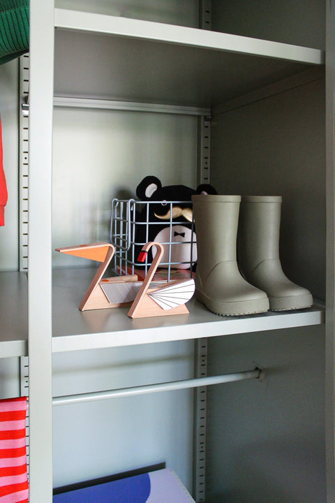 A pair of light gray rain boots displayed on a shelf next to a wire basket and decorative wooden pelican and swan, inside a gray storage closet.