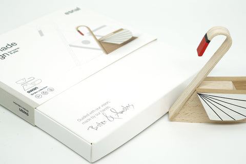 A wooden swan-shaped object with a red-tipped neck and white fan-like tail details stands beside its white product box on a clean white background.