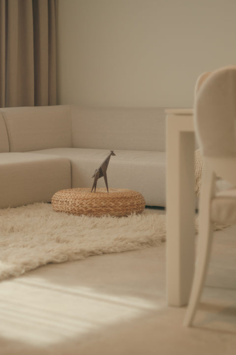 A styled interior scene with a cream sectional sofa, a fluffy cream rug, a round woven ottoman, and a small dark giraffe figurine standing on top. A white table edge and chair are visible in the foreground, with soft natural light across the room.