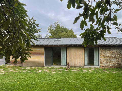 A modern single-story house with horizontal wood siding, a dark metal roof, and stone wall sections, set in a grassy yard with tree branches framing the top of the image.