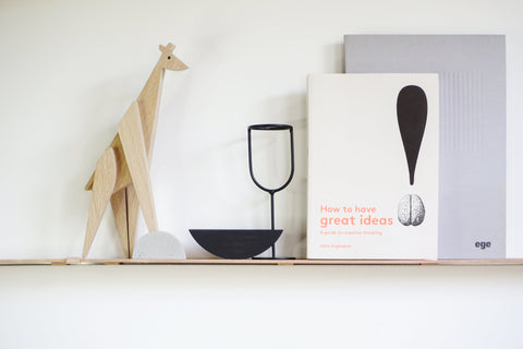 A styled interior shelf scene with a light wood giraffe figurine, a black bowl, a black metal stand, a white book titled "How to have great ideas," a gray notebook, and a gray folder against a white wall.