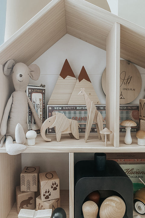 A styled wooden toy house shelf display featuring a plush mouse, natural wood animal figures, geometric mountain trees, wooden blocks, and a small black stove.