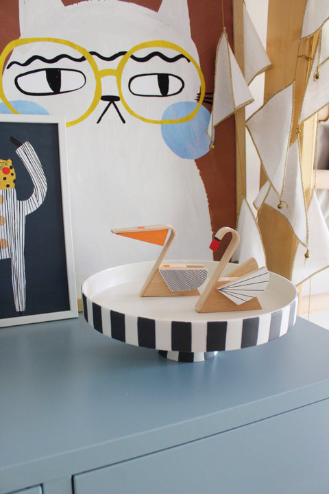 A decorative tray with black and white stripes holding two wooden figurines pelican and swan, styled on a blue cabinet in a room with whimsical wall art and hanging flags.