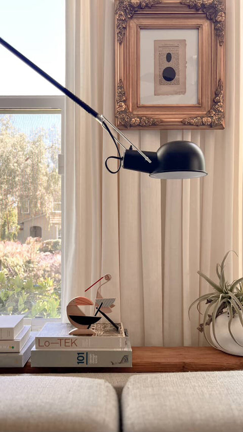 two wooden magnetic figurines around a modern black adjustable desk lamp with a dome shade and articulated arm, styled on a wooden table next to books and a small plant in a home office setting.